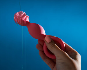 Woman pouring lubricant on pink anal beads on blue background.  - obrazy, fototapety, plakaty