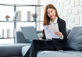 Asian professional successful happy female businesswoman secretary in formal business suit sitting smiling on cozy sofa holding laptop notebook computer and preparing paperwork document for meeting