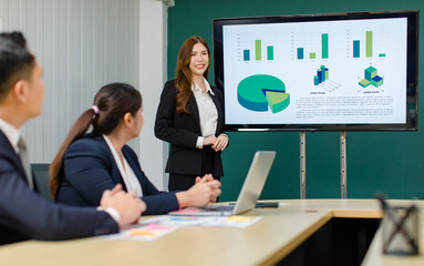 Asian professional successful smart confident female businesswoman presenter lecturer in formal business suit standing presenting graph chart document information on computer screen in meeting room
