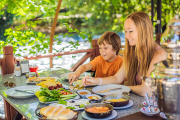 Mom and son eat Turkish breakfast. Turkish breakfast table. Pastries, vegetables, greens, olives, cheeses, fried eggs, spices, jams, honey, tea in copper pot and tulip glasses, wide composition