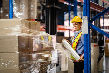 Female worker wrapped a parcel of stretch film on a cargo in a large factory warehouse. Logistics...