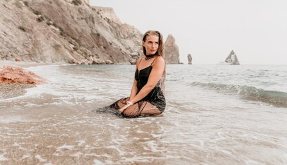Woman summer travel sea. Happy tourist in black dress enjoy taking picture outdoors for memories. Woman traveler posing on sea beach surrounded by volcanic mountains, sharing travel adventure journey