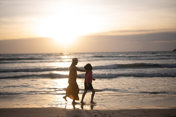 A young mother holds her little daughter by the hand stay holiday together along the beach towards the beautiful silhouette sunset. Happy mom and kid walking a long the white sand beach.