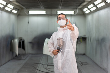 Portrait man in protective suit painting car with thump up in auto body shop