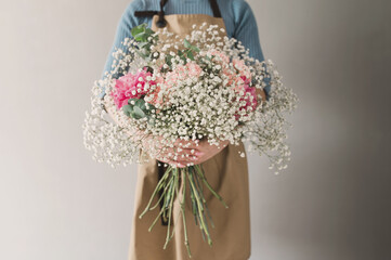 a young woman florist in an apron holds in her hands a ready-made delicate bouquet of gypsophila and pink peonies on a gray background close-up without a face. Banner. Impersonal