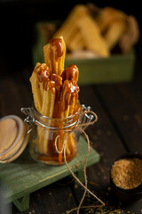 Churros served in a glass jar with caramel sauce, placed on a rustic wooden surface. Crispy golden pastry sticks, sugar crumbs, and warm tones create a cozy dessert atmosphere.