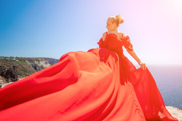 Fototapeta premium woman sea red dress. Blonde with long hair on a sunny seashore in a red flowing dress, back view, silk fabric waving in the wind. Against the backdrop of the blue sky and mountains on the seashore.