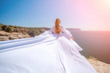 Fototapeta na wymiar woman sea white dress. Blonde with long hair on a sunny seashore in a white flowing dress, rear view, silk fabric waving in the wind. Against the backdrop of the blue sky and mountains on the seashore