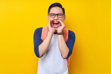 Excited young Asian boy student wearing casual clothes and backpack screams hot news about sales discounts with hands near mouth isolated on yellow background. high school university college concept