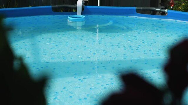 Light summer rain and sunshine over backyard pool. In the foreground, blurred leaves of flowers. Raindrops in the pool. The pool has a skinner installed to clean the water