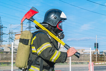 Naklejka premium A firefighter in a helmet and breathing apparatus put an assault axe on his shoulder. A firefighter in protective clothing and a mask on the side. A lifeguard with a fire axe. Rescue service.