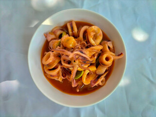 Indonesian menu of Cumi Asam Manis (sweet and sour squid or octopus) with pete on a white bowl. It...