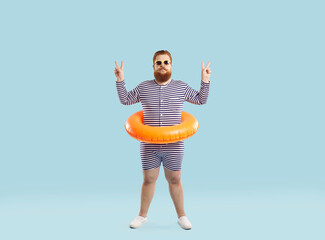 Funny chubby man with inflatable circle for swimming at waist shows V-sign on light blue...