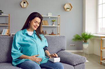 Happy pregnant woman measures blood pressure. Beautiful smiling young lady sitting on sofa,...