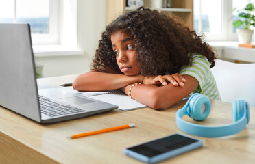 Tired curly African American girl teen bored with head on table in front of laptop sad and...