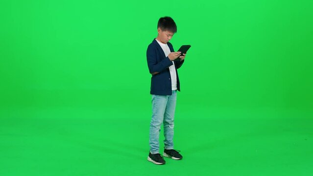 Asian schoolboy in a suit standing on a green background and using a screen tablet, reads an e-book, template on a chromakey.