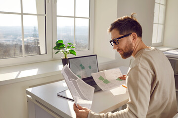 Portrait of serious focused attractive architect in process of work, sitting at desk at his workplace in office. Young engineer with glasses with cadastral maps and projects sitting in front of laptop - obrazy, fototapety, plakaty