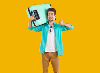 Portrait of happy excited man is showing thumb up carrying turquoise suitcase on shoulder going to vacations on yellow background. Banner for advertisement. Journey, tourism, travel, vacation concept.