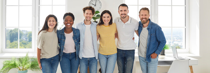 Group portrait of creative, smart and cheerful startup coworkers who are smiling at camera. Multiracial people in comfortable casual clothes hugging while standing in row in office. Panoramic banner.