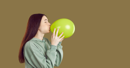 Woman blowing up balloon. Side view of young long haired brunette woman or teenage girl in casual green hoodie inflating yellow balloon on khaki copy space studio background. Birthday party concept