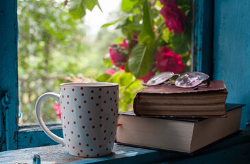 A cup, glasses and two books on the windowsill of an old wooden window, behind which roses are blooming
