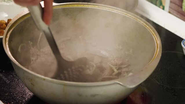 A woman's skimmer hand stirs the pieces of meat and onions frying in the cauldron. The liquid in the wok is boiling and steam is rising. Caucasian cuisine