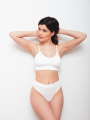 Fototapeta premium Portrait of young beautiful sexy woman in white lingerie. Smiling carefree model wearing pure underwear. Hot brunette posing near wall in studio interior. Perfect body. Slim and fit