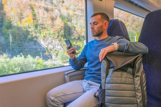 Male caucasian traveler rides in a suburban high-speed train with a backpack on the seat, prints a message in a smartphone phone looks out the window enjoying the trip.