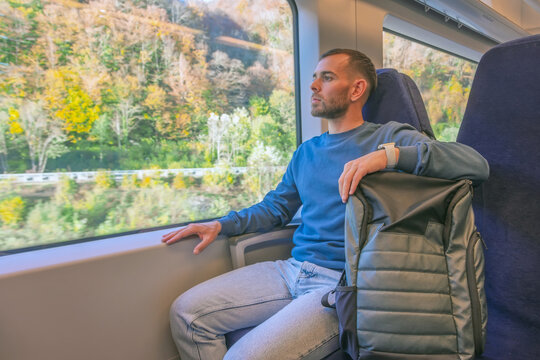 Male caucasian traveler rides in a suburban high-speed train with a backpack on the seat, looks out the window enjoying the trip.