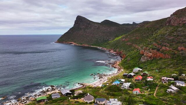 South Africa small beach town Smitswinkel Bay Cape of Good Hope Simon's Town aerial drone cinematic homes surf wave green lush spring summer deep aqua blue sea wind cloudy mountain backward motion