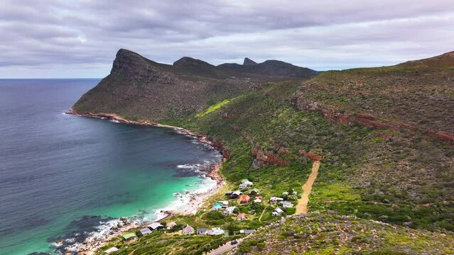 South Africa small beach town Smitswinkel Bay Cape of Good Hope Simon's Town aerial drone cinematic homes during green lush spring summer deep aqua blue ocean sea cloudy mountain cliffs forward motion