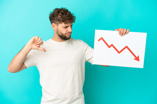 Young handsome caucasian man isolated on blue background holding a sign with a decreasing statistics arrow symbol and doing bad signal