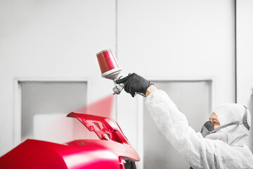 Worker paints auto with spray gun. Red bumper of car in a paint chamber during repair work. - 613784691