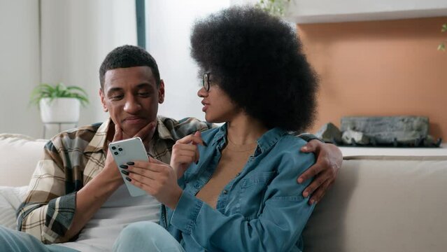 Happy family African American couple home couch sofa choosing products together shopping online discuss talking buying in internet shop app store smartphone ordering delivery purchase combined budget