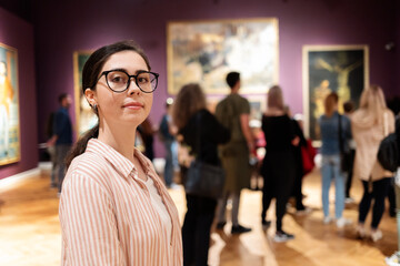 Portrait of young Caucasian woman wearing glasses. In background, people are looking at paintings. Concept of education and Museum Day