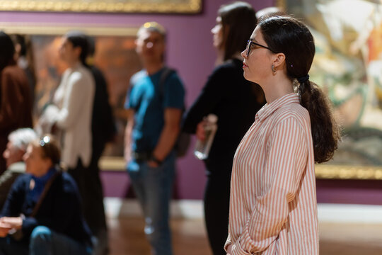 Side View Of Young Caucasian Woman Wearing Glasses And Looking At Exhibition. People And Masterpieces In Background. Concept Of Museum Day