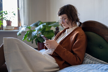 Focused pensive teenage girl using mobile phone while relaxing on bed at home, scrolling on social...