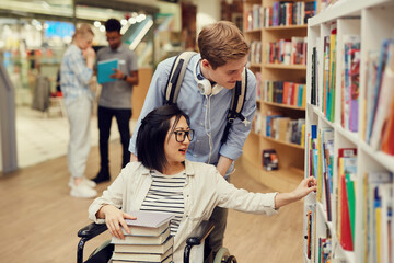 Careful young volunteer pushing wheelchair with Asian student girl while assisting her in library