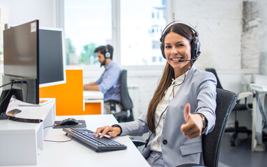 Happy customer service operator woman wearing headset, working on computer, and showing thumb up to...