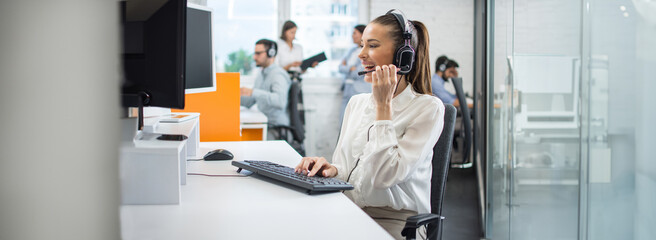 Smiling businesswoman using computer and listening to client via headset in call center office. Panoramic view.