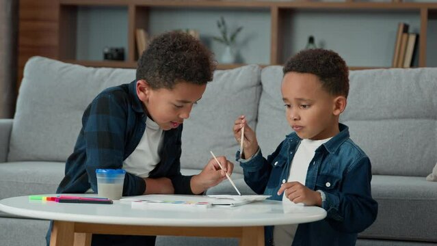 Two African American children kids school boys ethnic little friends siblings brothers painting together at home with colored watercolor paints picture in living room primary education family hobby