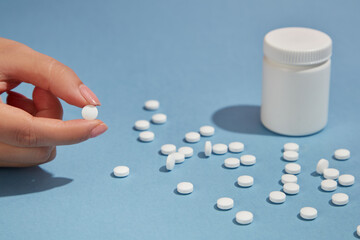 Fingers reach for a pill, several other round white pills with white bottle unbranded on a blue background. Minimal scene for advertising. Selective focus