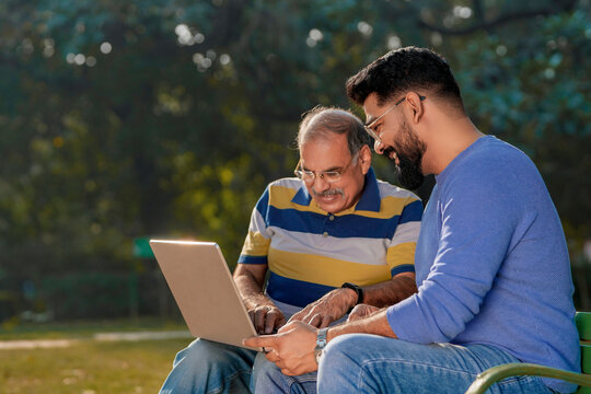 Young indian man showing bank card and laptop how to uses to his father at park
