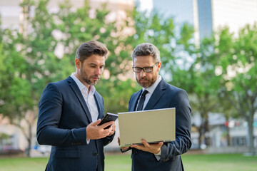 Two handsome young businessmen in classic suits using laptop. Handsome business man in suits working on laptop communication with business projects in city. Partners thinking and plan for business.