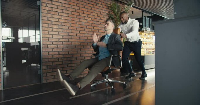 Cheerful leisure in the office. Two middle-aged men are having fun and one is carrying the other in an office chair