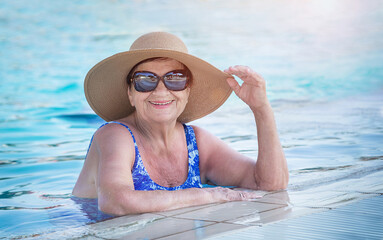 Senior woman (over age of 50) in swimsuit, hat and sunglasses relaxing in resort swimming pool. Happy retired female  enjoying summer vacation in hotel. All inclusive.