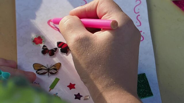 Close-up slowmotion shot of a child hand painting pink heart with marker on a greeting card. Small cute symbols: butterflies and curve lines. Hand-made present by a child