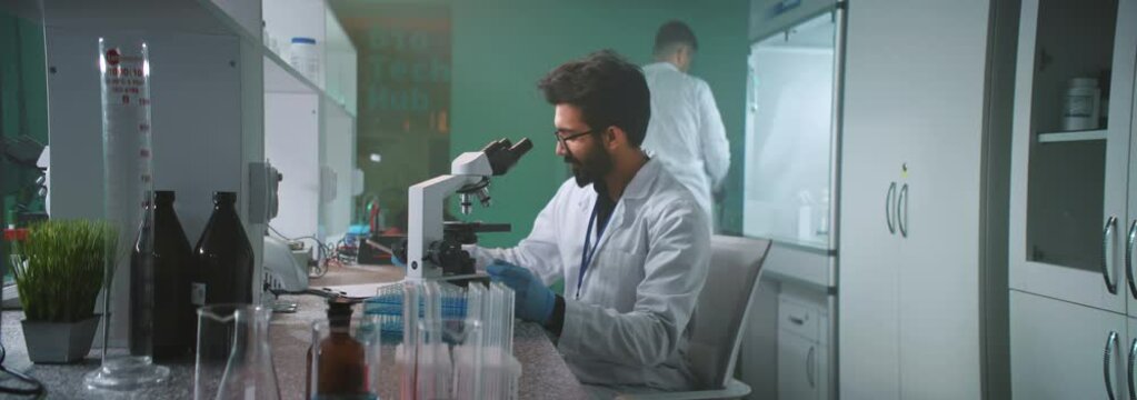 Slow motion young man scientist with glasses and protective gloves looking under the microscope holds a tube in the laboratory. Medicine, biotechnology, chemistry. On background colleague walking