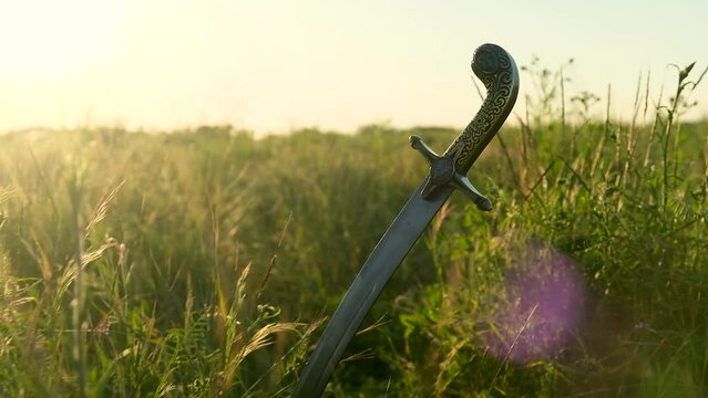 The saber of the Ukrainian Cossacks is stuck in the ground, among the grass, against the background of the sunset. Beautiful bokeh appears in the frame. The concept of freedom. Close-up.