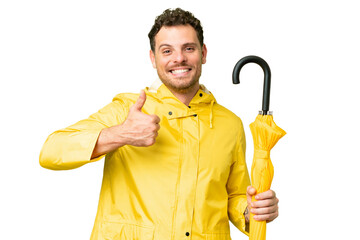 Brazilian man with rainproof coat and umbrella over isolated chroma key background giving a thumbs...